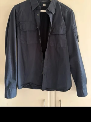  C.P. Company overshirt - Stilren mörkblå skjorta från C.P. Company med klassisk krage, två bröstfickor med lock och knappar framtill. Skjortan har lång ärm och är tillverkad i ett mjukt bomullstyg. Perfekt för en avslappnad men snygg look.