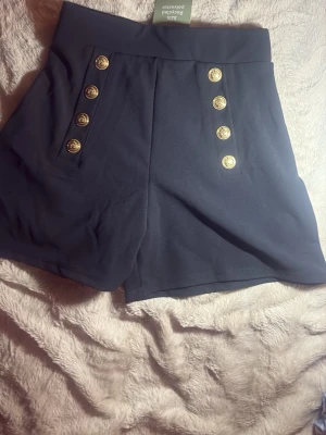 Svarta shorts med guldknappar - Stilrena svarta shorts med hög midja och sex dekorativa guldknappar framtill. Shortsen har en klassisk, något loose passform och är perfekta för en trendig look. Tillverkade i ett mjukt och bekvämt material.