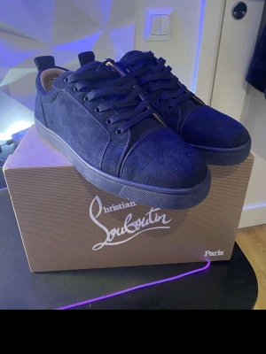 Mörkblå sneakers från Christian Louboutin - Snygga mörkblå sneakers från Christian Louboutin i mocka med klassiska röda sulor och diskret logga på sidan. Skorna har rund tå, svarta skosnören och låg profil som ger en clean och stilren look. Perfekta för dig som vill ha något exklusivt och trendigt.