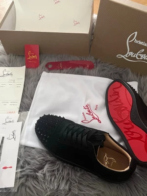 Svarta Louboutin sneakers med nitar - Snygga svarta sneakers från Christian Louboutin med ikonisk röd sula och svarta nitar på tån. Skorna har en låg profil och är tillverkade i mocka och läder. Perfekta för dig som vill sticka ut med exklusiv design och edgy detaljer. 