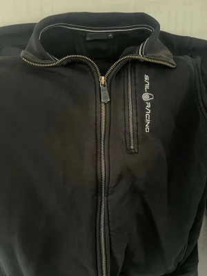 Svart ziptröja från Sail Racing - Svart ziptröja från Sail Racing med vit logga på bröstet och kontrastsömmar vid kragen. Tröjan har hel dragkedja och mjukt fleecefoder på insidan. Perfekt för en sportig och avslappnad stil.