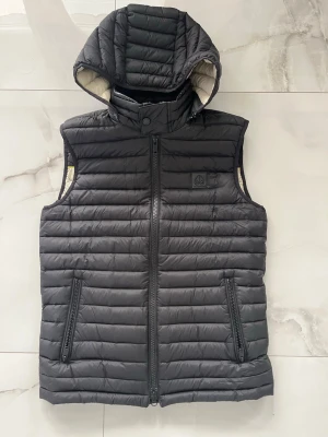 Svart dunväst med huva från Pyrenex - Snygg svart dunväst från Pyrenex med avtagbar huva och quiltad design. Västen har två dragkedjeförsedda fickor framtill och en hög krage med tryckknapp. Perfekt för lager-på-lager och streetstyle.