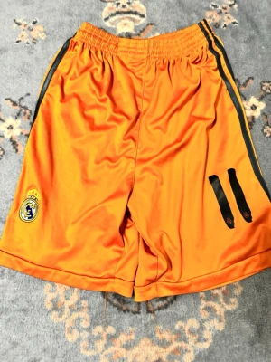 Orange Real Madrid shorts från Adidas - Snygga orange shorts från Adidas med Real Madrid-logga på ena benet och svarta detaljer längs sidorna. Tillverkade i ett lätt och sportigt syntetmaterial, perfekta för träning eller fotboll. Elastisk midja för skön passform och klassisk Adidas-design.