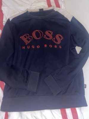 Mörkblå sweatshirt från Hugo Boss - Mörkblå sweatshirt med stora röda BOSS-bokstäver och HUGO BOSS-tryck på bröstet. Tröjan har långa ärmar, ribbade muddar och kontrastdetaljer vid axlarna. Perfekt för en avslappnad och trendig stil.