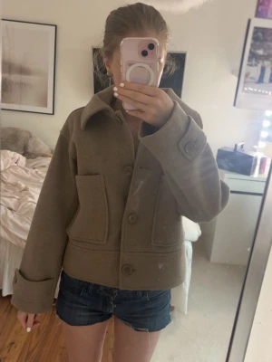 croppad jacka från Kappahl". - Stilren beige jacka från Kappahl i ullblandning. Jackan har stora framfickor, klassisk krage och knäppning med stora knappar framtill. Croppad modell med lång ärm och enkel, clean design som passar till många outfits.