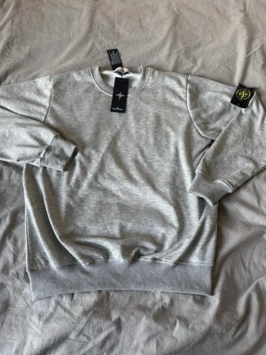 Stone Island Sweatshirt  - Snygg grå sweatshirt från Stone Island med klassisk rund hals och ribbade muddar. Tröjan har den ikoniska svarta patchen med gul och grön logga på höger ärm. Perfekt för en clean och stilren look.