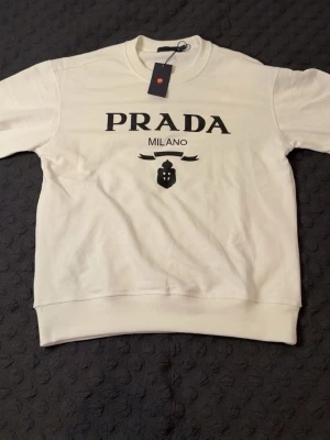 Vit sweatshirt från Prada Milano - Snygg vit sweatshirt från Prada Milano med svart logotyp och emblem på bröstet. Tröjan har rund halsringning och ribbade muddar vid ärmslut och nederkant. Perfekt för dig som gillar exklusiva märken och stilren design. STORLEK M, PASSAR M OCH L. HELT NY, OANVÄND.