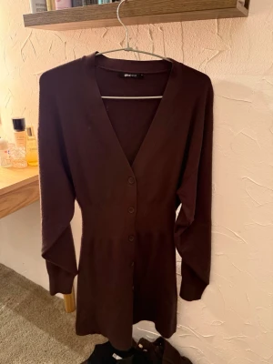 Brun lång klänning från Gina Tricot - Mysig lång brun klänning från Gina Tricot med v-ringning och knäppning framtill. Klänningen har långa ärmar och en rak passform.