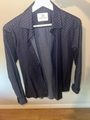 Mönstrad blå skjorta The Shirt Factory - Snygg blå skjorta från The Shirt Factory med små diskreta mönster i hela tyget. Skjortan har klassisk krage, långa ärmar och knäppning framtill. Perfekt för dig som vill ha en stilren men ändå unik look. Aldrig använd!