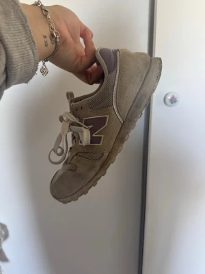 Beige New Balance sneakers med lila detaljer - Säljer ett par beigea New Balance sneakers med lila detaljer och klassisk N-logga på sidan. Skorna har snörning, meshpaneler för extra andningsförmåga och mockadetaljer. Perfekta för dig som gillar en clean och sportig stil.