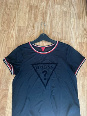 Svart topp från Guess - Snygg svart topp från Guess med klassisk logga framtill. T-shirten har korta ärmar och ribbade kanter med röd, vit och svart rand. Då den är ”genomskinlig” är den en perfekt lager på lager topp.