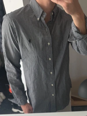 Svartvit rutig skjorta från Ralph Lauren - Klassisk svartvit rutig skjorta från Ralph Lauren med button-down krage och broderad logga på bröstet. Skjortan har långa ärmar och vita knappar framtill. Perfekt för en stilren look.