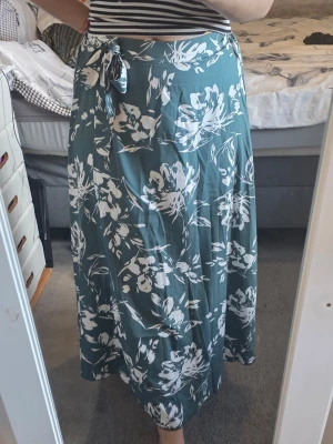 Grön blommig omlottkjol med knyt - Snygg långkjol i grönt med vitt blommönster och omlottmodell. Kjolen har en knytning i midjan som ger en trendig och avslappnad look. Perfekt för dig som gillar en flowy och somrig stil. Storlek M