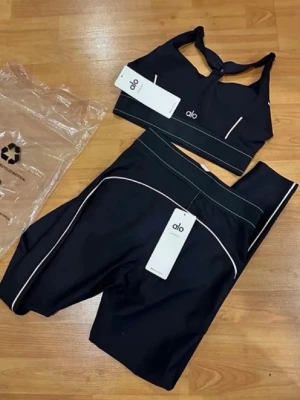 Svart träningsset från Alo - Snyggt svart träningsset från Alo med sport-bh och höga tights. Setet har vita kontrastsömmar och är gjort i ett stretchigt material som passar perfekt för träning. Sport-bhn har en twistad detalj fram och tightsen har bred midjeresår.