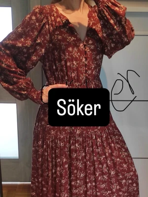 Blommig vinröd långklänning - Romantisk vinröd långklänning med småblommigt mönster och långa puffärmar. Klänningen har markerad midja, v-ringning och knappar framtill. Perfekt för dig som gillar bohemisk stil och mjuka material.