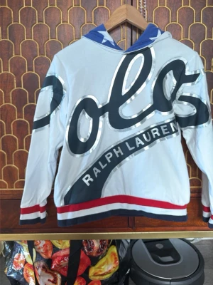 Vit hoodie från Polo Ralph Lauren - Snygg vit hoodie från Polo Ralph Lauren med stor logga i svart och silver över hela framsidan. Huva med blå insida och vita stjärnor, samt röda och marinblå ränder vid ärmslut och nederkant. Perfekt för dig som gillar streetwear och vill sticka ut.