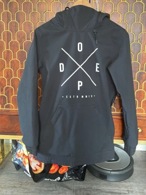 Svart vindjacka från Dope - Snygg svart vindjacka från Dope med stor vit logga och text på ryggen. Jackan har huva med dragsko, magficka och elastiska muddar vid ärmsluten. Perfekt för dig som vill ha en stilren och streetinspirerad look.