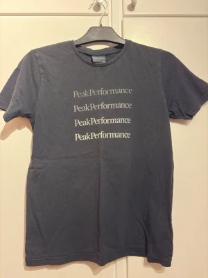 Mörkgrå Peak Performance t-shirt - Snygg mörkgrå t-shirt från Peak Performance med tryckt logga i vitt på bröstet. Klassisk rund hals och korta ärmar. Perfekt för en avslappnad och stilren look. Tillverkad i mjuk bomull som känns skön mot huden.