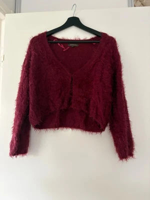 Vinröd fluffig kofta Pixy Cardigan - Vinröd fluffig kofta med v-ringning och lång ärm. Modellen är croppad och har en mjuk, lurvig känsla. Tillverkad i 70% polyamid och 30% akryl. Perfekt för dig som vill ha en mysig och trendig look.