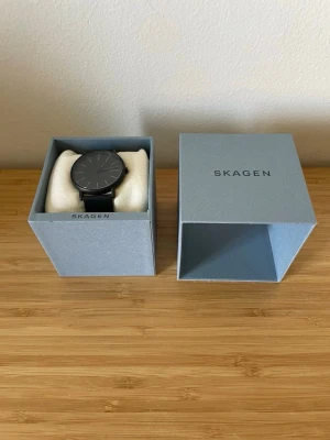 Svart minimalistisk klocka från Skagen - Stilren svart klocka från Skagen med rund urtavla och svart läderarmband. Klockan har en minimalistisk design med tunna visare och diskreta markeringar. Perfekt accessoar för en modern look.