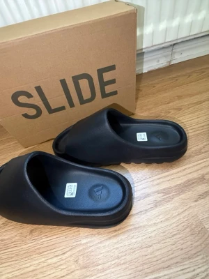 Svarta Adidas Yeezy Slide tofflor - Säljer ett par svarta Adidas Yeezy Slide tofflor med stilren och chunky design. Tillverkade i mjukt och flexibelt syntetmaterial för maximal komfort. Perfekta för att chilla hemma eller dra ut snabbt. Klassisk Adidas-logga i sulan och boxen medföljer. Oanvända pris kan diskuteras. Fråga gärna om fler bilder.
