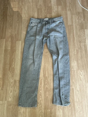 Grå raka jack&jones jeans med rå kant Relaxed/Chris fit - Säljer ett par grå jeans med rak passform och råa kanter nertill. Klassisk femficksmodell med dragkedjegylf och bälteshällor. Jeansen har en neutral grå tvätt och är tillverkade i denim. Perfekta för en avslappnad och trendig look.