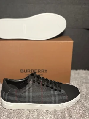Burberry  sneakers - Snygga sneakers från Burberry i svart och grått med klassiskt rutmönster och diskreta röda linjer. Skorna har vita sulor, svarta skosnören och detaljer i läder vid häl och tå. Perfekta för dig som gillar stilrena och exklusiva sneakers.