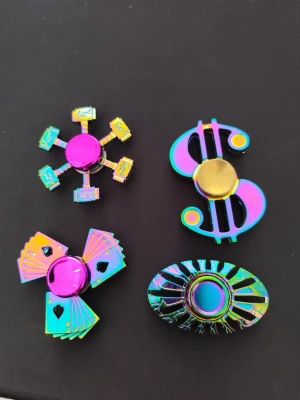 Fidget spinners i regnbågsfärgat metall - Fyra unika fidget spinners i regnbågsfärgat metall, med former som dollartecken, spelkort, pokerchips och kasinotema. Perfekta som samlarprylar eller för att hålla händerna sysselsatta. Säljs som set.
