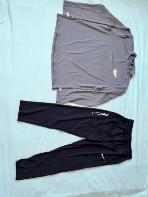 Nike träningsset grå tröja & svarta byxor - Nike träningsset med en grå långärmad tröja med halv dragkedja och reflexdetaljer samt svarta träningsbyxor med resår i midjan och Nike-logga. Perfekt för löpning eller gym. Materialet är lätt och andas, vilket gör det bekvämt under träning.