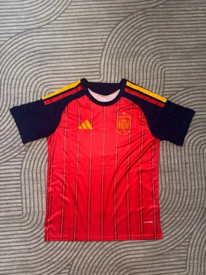 Spaniens fotbolls tröja - Röd och marinblå fotbollströja från Adidas med Spaniens landslagsemblem på bröstet och gula detaljer på axlarna. Tröjan har korta ärmar och är tillverkad i ett lätt och ventilerande material, perfekt för träning eller match.