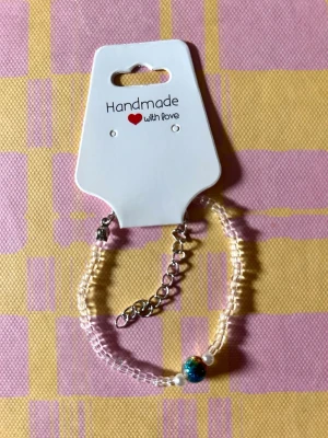 Handgjort pärlarmband med färgglad detalj - Snyggt handgjort armband med små vita pärlor och en större glittrig pärla i blått och grönt som mittpunkt. Armbandet har justerbar silverfärgad kedja och passar perfekt för att ge din stil en unik touch.