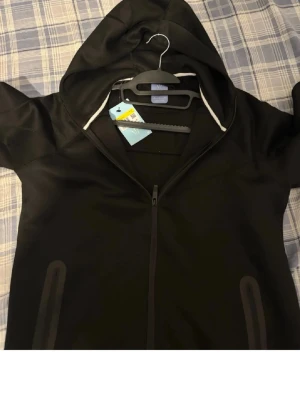 Svart hoodie från Nike NOCTA - Svart hoodie från Nike NOCTA med dragkedja framtill och två fickor. Hoodien har en huva med vita snören och är tillverkad i ett mjukt material som känns skönt mot huden. Perfekt för en avslappnad streetwear-look.
