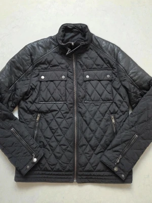Zara Man, Svart quiltad pufferjacka med detaljer - Snygg zara Man svart pufferjacka med quiltat mönster och coola skinndetaljer på axlar och ärmar. Jackan har två bröstfickor med knappar, dragkedjor vid ärmslut och ståkrage med spänne. Perfekt för dig som vill ha en stilren och trendig jacka. Size M.