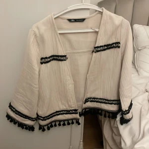 Beige kofta med svarta detaljer från Zara - Säljer en beige kofta från Zara med svarta broderade band och fransar längs ärmar och nederkant. Koftan har en avslappnad passform, knytband framtill och är tillverkad i ett mjukt bomullstyg. Perfekt för dig som gillar boho-vibe och snygga detaljer. Den är i storlek XS och passar mig som har S.