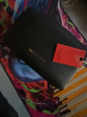 Svart handväska från Valentino - Snygg svart handväska från Valentino med stilren design och guldfärgad logga framtill. Väskan är tillverkad i ett strukturerat material och har en praktisk ficka på baksidan. Perfekt accessoar för att lyfta din outfit.