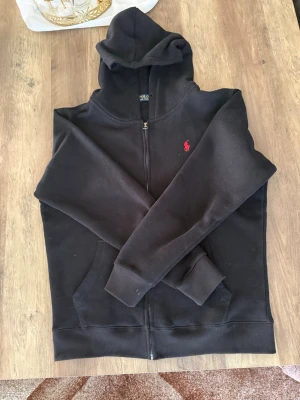 Svart hoodie från Polo Ralph Lauren - Svart hoodie med dragkedja från Polo Ralph Lauren. Klassisk design med huva, kängurufickor och röd broderad logga på bröstet. Perfekt för en chill och stilren look. Tillverkad i mjukt bomullsmaterial för extra komfort.