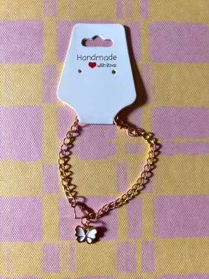 Guldfärgat armband med fjäril - Snyggt guldfärgat armband med en vit emaljerad fjärilsberlock. Länkarna är mellanstora och armbandet har en trendig kedjedesign. Perfekt accessoar för att lyfta din outfit med en söt detalj.