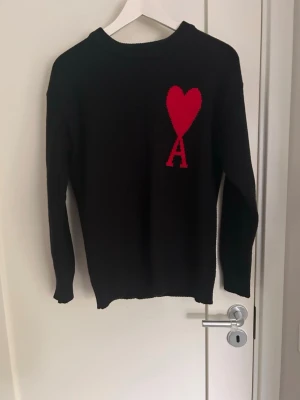 Svart stickad tröja från Ami - Svart stickad tröja från Ami med rund halsringning och ribbade muddar. På bröstet finns ett stort rött hjärta med bokstaven A, vilket är signaturdetaljen från märket. Perfekt för dig som gillar stilrena plagg med en twist.
