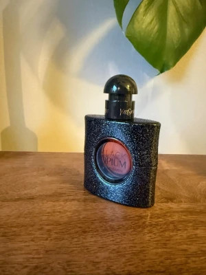Yves Saint Laurent Black Opium parfym - Säljer en ikonisk Black Opium parfym från Yves Saint Laurent. Flaskan är svart med glittrig yta och har en rund öppning i mitten där parfymens namn syns. Korken är svart och rundad. Perfekt för dig som gillar lyxiga och djärva dofter.