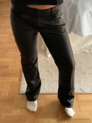 Svarta bootcut jeans i skinnlook - Säljer ett par svarta bootcut jeans med skinnliknande yta. Byxorna har klassisk femficksdesign och normal midja. Snygg passform som sitter tajt upptill och släpper vid benen. Perfekta för dig som vill ha en edgy stil.