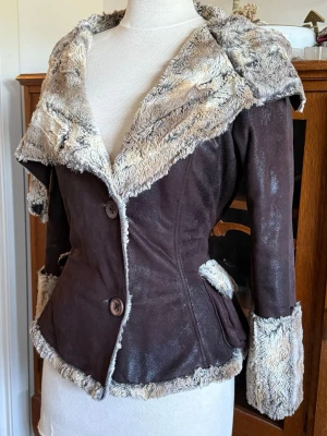 Brun pälsjacka med fuskpälsdetaljer - Snygg brun jacka med stora fuskpälsdetaljer i beige och grå toner på krage, ärmslut och nederkant. Figursydd modell med två knappar framtill och fickor på sidorna. Perfekt för dig som vill ha en varm och trendig vinterlook. 