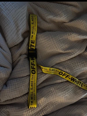 Gult Off-White bälte med svart text - Säljer ett ikoniskt gult bälte från Off-White med svart text och metallspänne. Bältet har den klassiska industriella looken med tryckt text 'Weight Securing System' och '5,400 lbs'. Perfekt för att lyfta din outfit med streetwear-vibe.