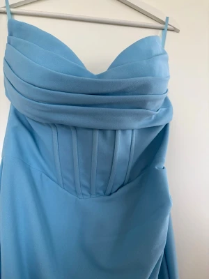 Ljusblå långklänning med draperad byst - Säljer en elegant ljusblå långklänning med smala axelband och draperad byst. Klänningen har en figurnära passform upptill och faller snyggt nedtill. Materialet känns som polyester eller satin och modellen har en lätt slits framtill.
