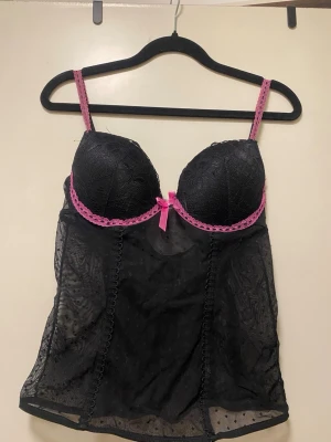 Svart spetsbody med rosa detaljer - Snygg svart body i mesh och spets med vadderade kupor, rosa spetskant och en söt rosa rosett framtill. Justerbara axelband i rosa. Perfekt till fest!