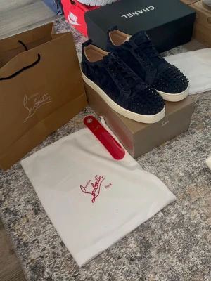 Mörkblå Louboutin sneakers med nitar - Snygga mörkblå sneakers från Christian Louboutin i mocka med svarta nitar på tån och matchande mörkblå skosnören. Skorna har en vit sula som kontrast och klassisk låg siluett. Perfekta för dig som vill sticka ut med exklusiv stil.