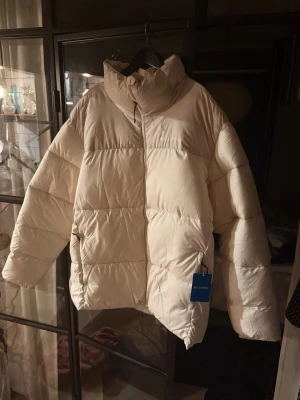 Vit pufferjacka från Columbia - ny med prislapp  - Säljer en riktigt fet vit pufferjacka från Columbia med hög krage och dragkedja framtill. Jackan är tjock och quiltad, perfekt för kalla vinterdagar. Materialet är syntet och jackan har två sidofickor med dragkedja. Passar dig som vill ha en oversized look. Ny med prislapp!