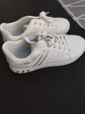 Vita sneakers från SSNJ med silverdetaljer - Stilrena vita sneakers från SSNJ med silvriga ränder och textdetaljer på sidan. Skorna har klassisk låg modell, vita snören och diskret mönstrad sula. Tillverkade i syntetmaterial med bekväm innersula och enkel design.