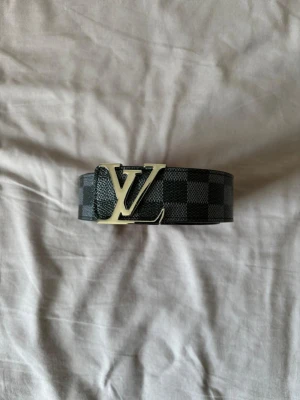 Louis Vuitton bälte - Snyggt svart bälte från Louis Vuitton med klassiskt rutigt mönster och ikoniskt LV-spänne i silver. Bältet är brett och har en lyxig känsla, perfekt för att lyfta vilken outfit som helst.
