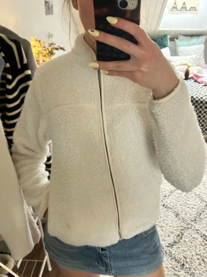 Vit fleece med dragkedja - Mysig vit fleece med hög krage och hel dragkedja framtill. Jackan har en fluffig, mjuk struktur och raka ärmar. Perfekt för att hålla värmen och ge en chill look. Passar till jeans eller kjol för en avslappnad stil.