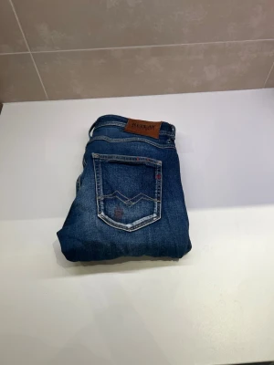 Blå jeans från Replay - Säljer ett par snygga blå jeans från Replay med klassisk femficksdesign och tydliga slitningar. Jeansen har en läderpatch med logga bak och detaljerade sömmar. Perfekta för dig som gillar en avslappnad och trendig stil. Har en liten fläck där bak men som inte syns särskilt mycket när man bär dem.